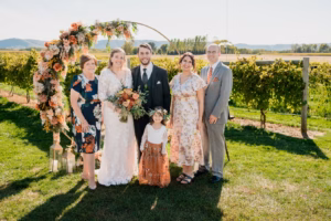 Six people, including a bride and groom, stand smiling outdoors in front of a floral wedding arch, with greenery and fields in the background.