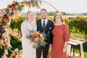 Three people stand outdoors in front of a floral wedding arch and vineyard. The woman on the left wears a white gown, the man in the middle a suit, and the woman on the right a red dress.