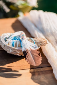 A brown and white feather rests on a shell bowl containing wrapped sage, placed on a wooden surface next to a light-colored cloth.
