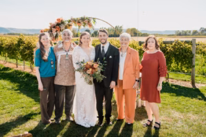 Bride and groom pose outdoors with four older adults in front of a floral wedding arch and vineyard on a sunny day.