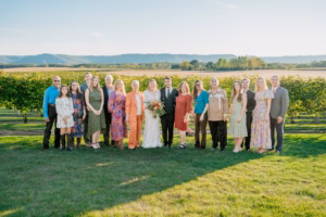 A group of people poses for a photo outdoors in a vineyard, with a couple dressed as bride and groom at the center. The group is dressed in semi-formal attire under clear skies.