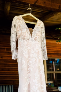 Long-sleeve white lace wedding dress with floral patterns hanging on a hanger against a wooden wall background.