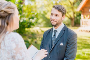 A man in a suit stands outdoors, facing a woman in a white dress who is holding a book, with greenery and a wooden structure in the background.
