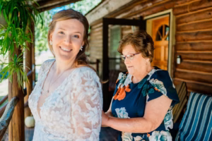 A woman in a floral dress fastens the back of a bride’s lace gown on a porch with wooden walls and greenery in the background.