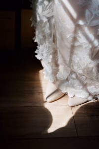Close-up of a person wearing a white lace dress and cream-colored pointed shoes standing on a wooden floor with sunlight casting shadows.