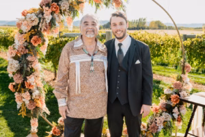 Two men posing and smiling in front of a circular floral arch outdoors, one in a patterned shirt with bolo tie, the other in a black suit and tie. Vineyards are in the background.