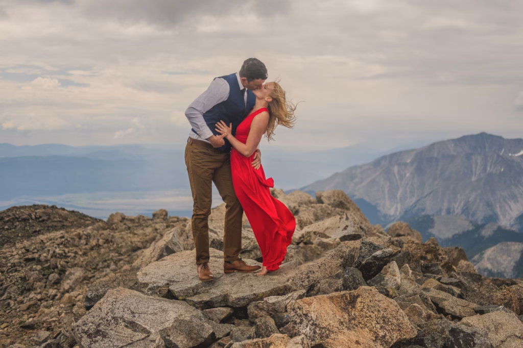 A man and woman stand on rocky terrain atop a mountain; the man dips the woman, who wears a red dress, as they kiss with mountains and clouds in the background.