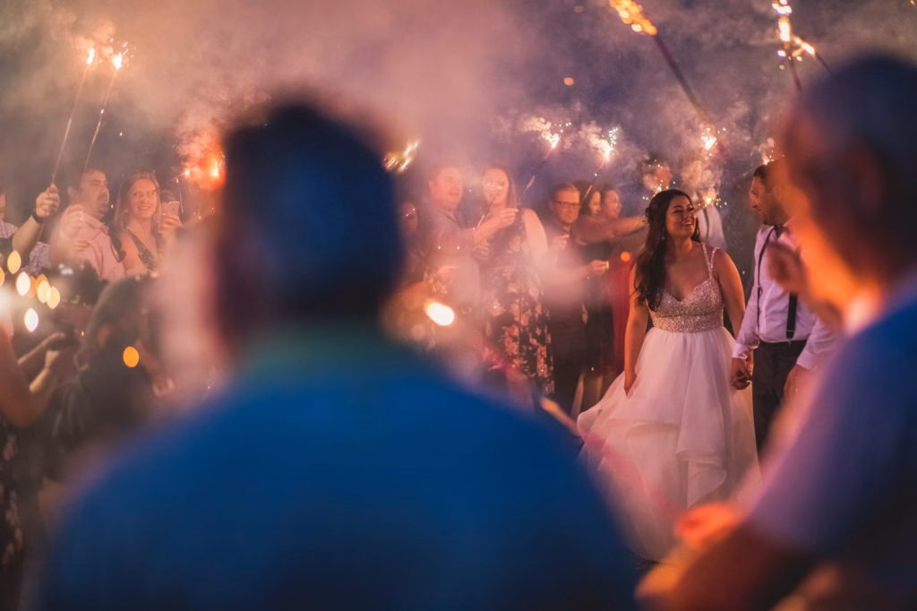 A bride and groom walk hand in hand through a crowd holding sparklers, with smoke and people blurred in the foreground.