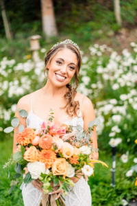 A woman in a white dress and tiara stands outdoors holding a large bouquet of colorful flowers, smiling at the camera with a background of greenery—captured beautifully by a Vail Wedding Photographer.