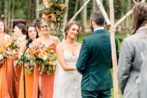 A bride and groom stand facing each other at an outdoor ceremony, surrounded by bridesmaids in shades of orange, as a Vail Wedding Photographer captures every cherished moment.