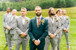 A groom in a dark green suit stands before five groomsmen in light gray suits, posing on grass with trees behind them—captured by a Vail Wedding Photographer.