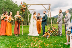 A bride and groom stand under a floral wedding arch with bridesmaids, groomsmen, and their dog on a grassy outdoor setting surrounded by trees, beautifully captured by a Vail Wedding Photographer.