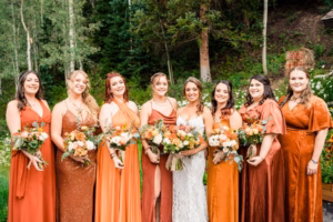 Eight women in coordinated orange-toned dresses and one in a white gown, all holding bouquets, stand outdoors in a lush, wooded area—captured beautifully by a Vail wedding photographer.