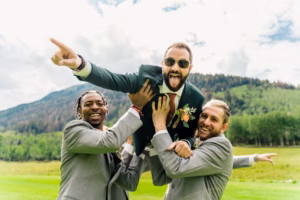 Two men in grey suits lift a man in a green suit and sunglasses, who points and sticks out his tongue, while a Vail Wedding Photographer captures the fun moment against a scenic backdrop of trees and mountains.
