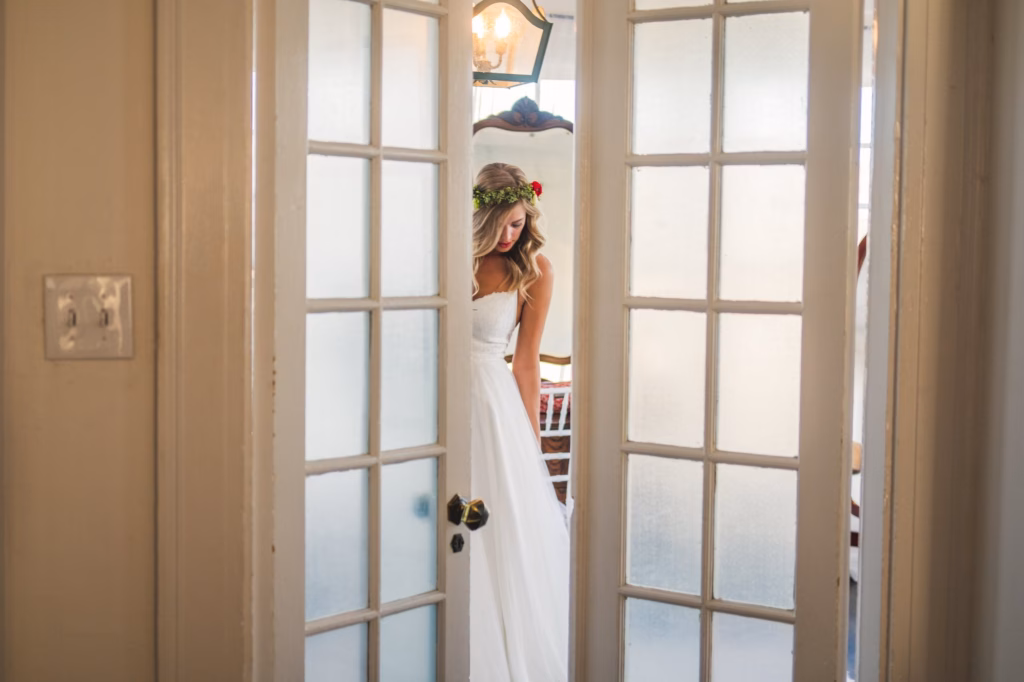 A woman in a white wedding dress and floral headpiece stands behind partially opened glass doors, looking downward.