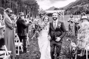 A bride and groom walk down the aisle outdoors, smiling and holding hands as their dog trots beside them. Captured by a Vail Wedding Photographer, this festive moment is surrounded by joyful guests amid stunning mountain scenery.