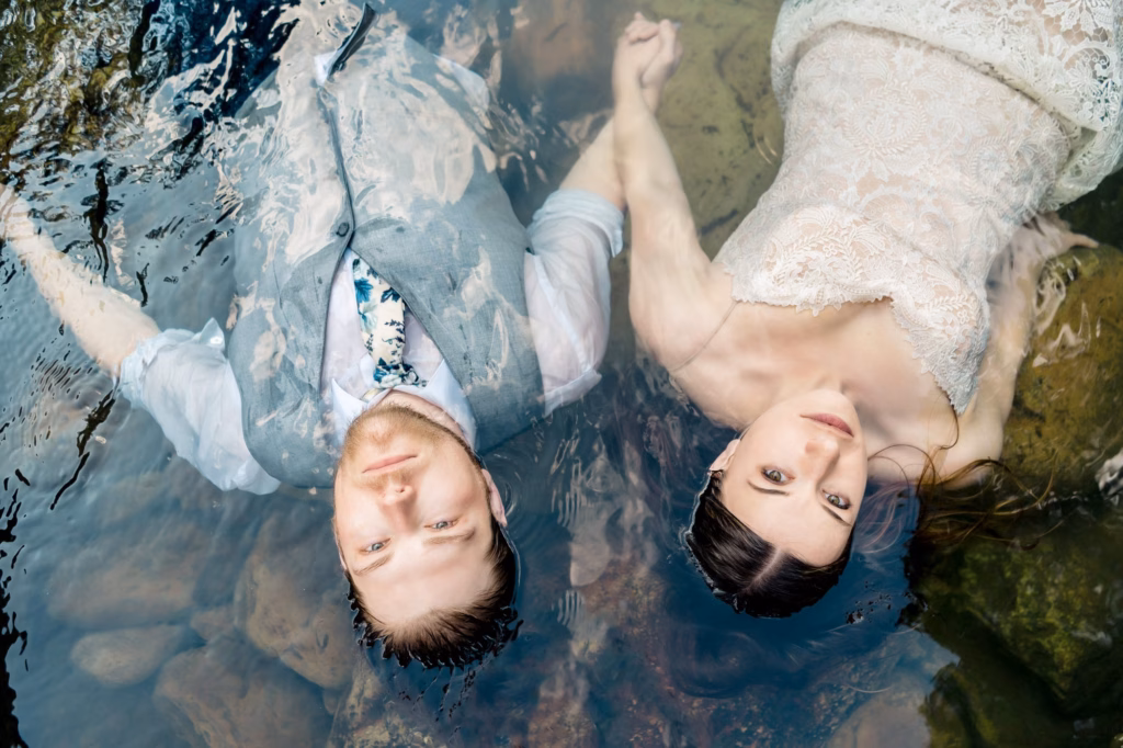 A man in a vest and tie and a woman in a lace dress lie side by side, holding hands, partially submerged in clear water with rocks beneath.
