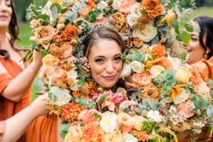 A woman wearing a tiara is surrounded by a large arrangement of colorful flowers, with several people holding the bouquet around her face—captured artfully by a Vail Wedding Photographer.