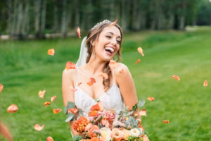 A bride in a white dress holding a bouquet smiles as flower petals fall around her on a grassy lawn, beautifully captured by a Vail Wedding Photographer.
