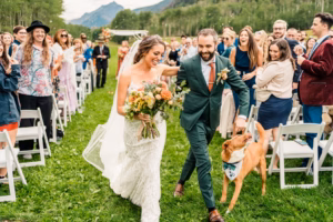 A bride and groom walk down an outdoor aisle with their dog on a leash, surrounded by wedding guests and breathtaking mountain scenery—perfectly captured by a Vail Wedding Photographer.
