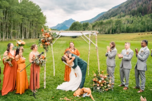 A bride and groom kiss under a rustic arch at an outdoor ceremony, surrounded by their wedding party in orange and gray attire, as a Vail Wedding Photographer captures the moment with mountains and trees in the background.