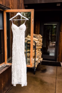 A white lace wedding dress hangs on a hanger from an open window, with a stack of firewood and a wooden building in the background—captured perfectly by a Vail Wedding Photographer.