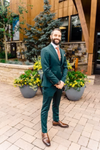 A man in a green suit with brown shoes and tie stands smiling on a stone patio, with potted flowers and a tree in the background—captured perfectly by a Vail Wedding Photographer.