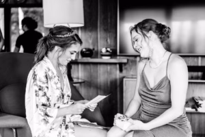 Two women sit indoors; one in a robe reads a card while smiling, and the other in a dress sits beside her, both captured candidly by a Vail Wedding Photographer in this relaxed, joyful moment.