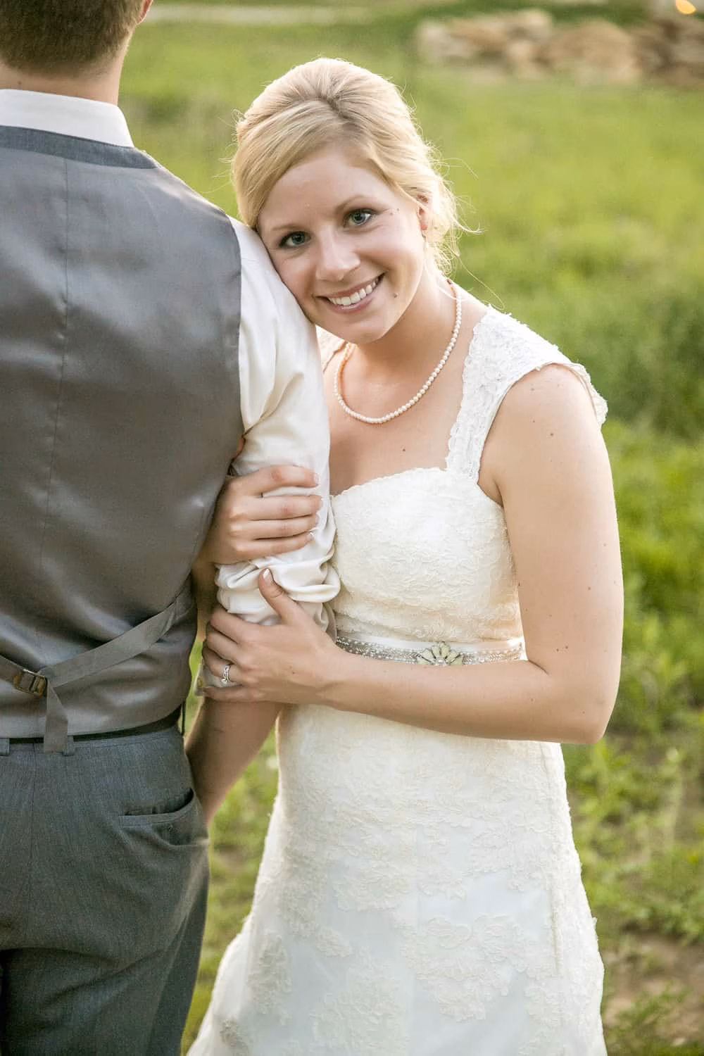 A bride in a white lace dress smiles while holding the arm of a man in a vest and dress shirt, standing outdoors on grass.