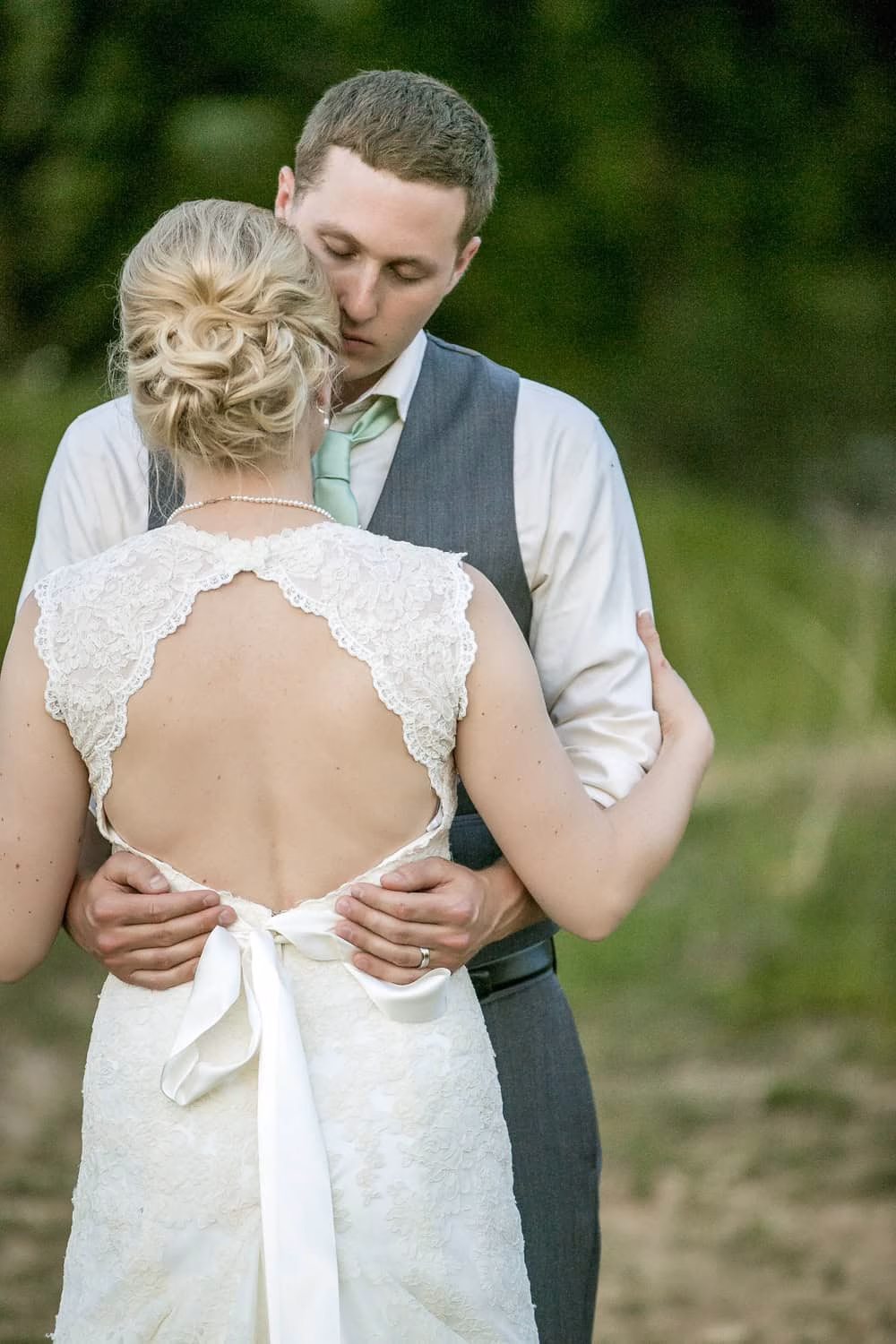A couple embraces outdoors; the woman wears a white lace-back dress and the man wears a vest and tie. They appear to be at a wedding.