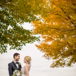 A bride and groom stand outdoors under green and yellow autumn trees, facing each other and smiling, with the bride holding a bouquet.
