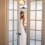 A woman in a white dress and floral headpiece stands between partially open glass doors in a softly lit room.