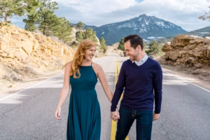 A woman in a teal dress and a man in a navy sweater hold hands and walk down a mountain road, with rocky hills and a snow-capped peak in the background.