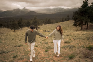 A man and woman holding hands walk through a grassy field with mountains and pine trees in the background under a cloudy sky.