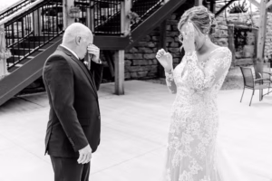 A bride in a lace dress and an older man in a suit wipe their eyes while standing outside near a staircase.