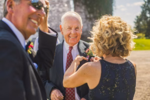 An older man in a suit and red tie smiles as a woman affixes a boutonniere to his jacket outdoors, with another man in sunglasses nearby.