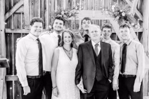 A group of six men and one woman, dressed in formal attire, pose together in front of a wooden backdrop decorated with flowers.