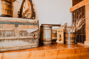A rustic scene with a large vintage trunk, weathered barrels, an old-fashioned phone, a mirror, and dried plants on a wooden surface near a staircase.