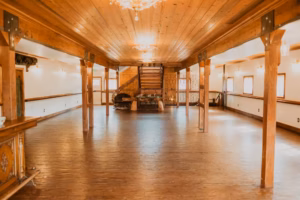 Spacious wooden hall with polished floors, exposed beams, warm lighting, chandeliers, and a staircase at the far end.
