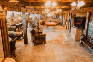 A rustic basement game room features wooden beams, leather chairs, a pool table, stone walls, and warm overhead lighting.