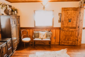 A rustic room with wooden walls and floor, a wooden bench with decorative pillows, a chair, an armoire, and a wooden door, illuminated by natural light from a window.