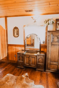 A rustic wooden vanity with a mirror sits against a wood-paneled wall, next to a tall cabinet and a framed vintage photo; animal hide rug on wooden floor.