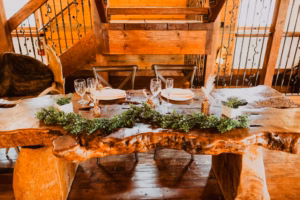 A rustic wooden dining table set with plates, glasses, and cutlery, decorated with greenery and small potted plants, in a wooden interior setting.