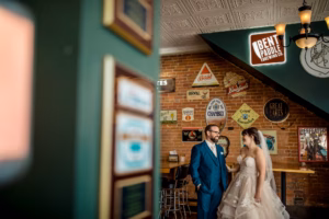A bride and groom stand together inside a bar decorated with vintage beer signs and brick walls, with a counter and barstools in the background.
