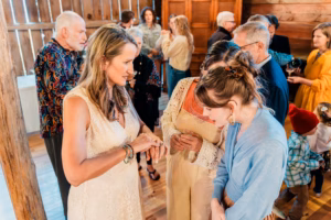 A woman in a white dress shows her ring to two women in a crowded indoor gathering with wooden walls and several people conversing in the background.