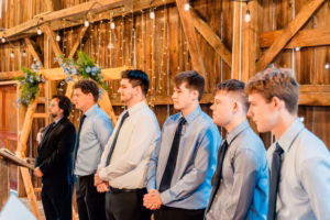 Five men in dress shirts and ties stand in a row next to an officiant in a barn decorated with string lights and floral arrangements.