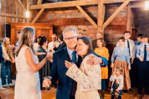 A smiling man in a suit embraces a woman in a white lace top at an indoor event, with a group of people gathered in the background.