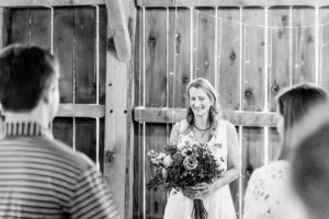 A woman in a white dress holding a bouquet stands indoors, facing two people. She is smiling, with wooden walls and string lights in the background.