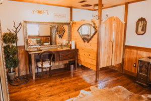 A rustic room with wood floors, a vanity table and chair, wall mirror, tree-shaped coat rack, and a wooden partition separating the space.