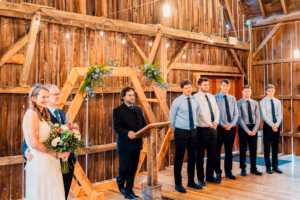 A bride holding a bouquet stands next to a man and a person at a podium, with six men in dress shirts and ties standing in a row inside a rustic wooden venue.